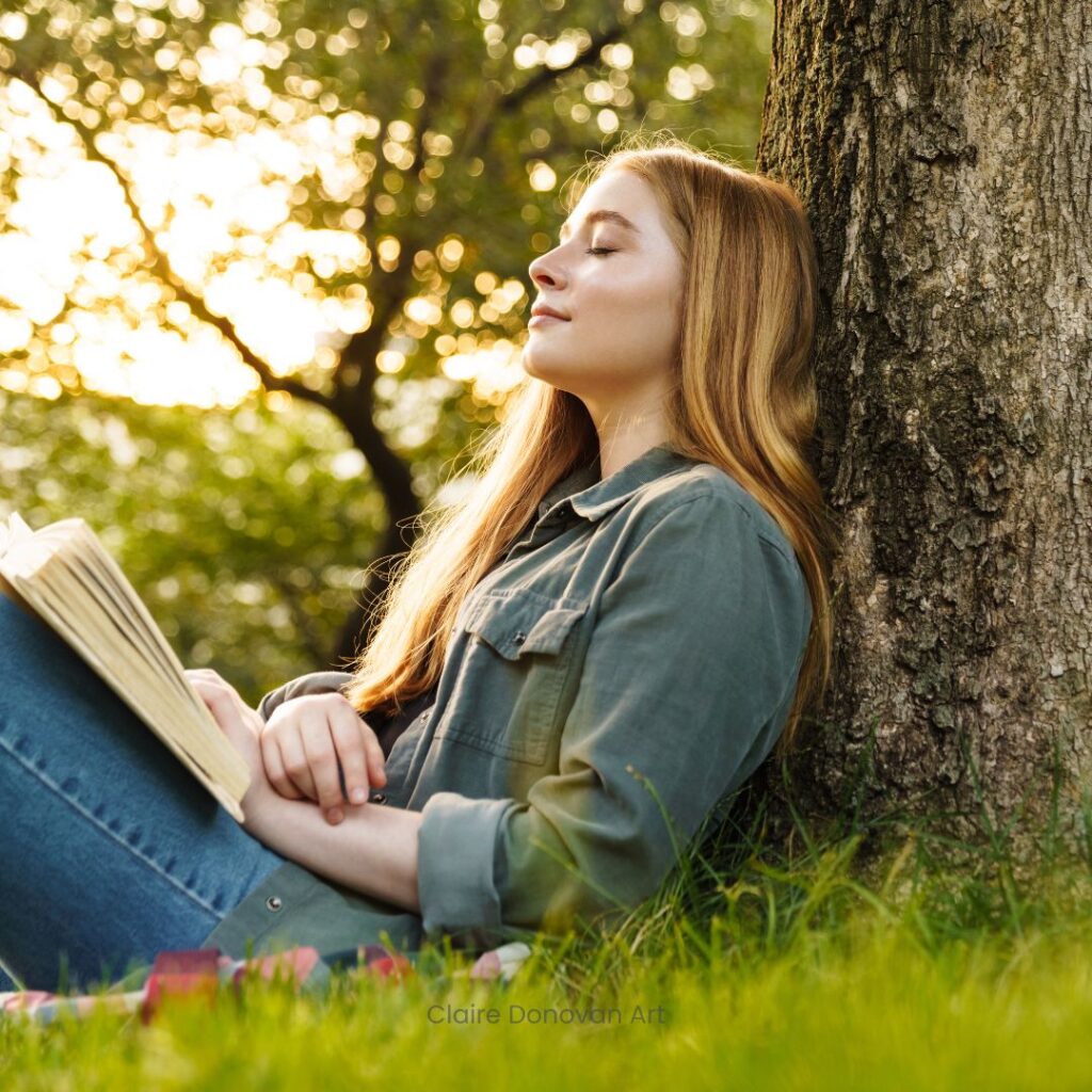 Mother Nature - being creative outside. A strawberry blonde woman leaning against a tree with her eyes closed and a book in her lap